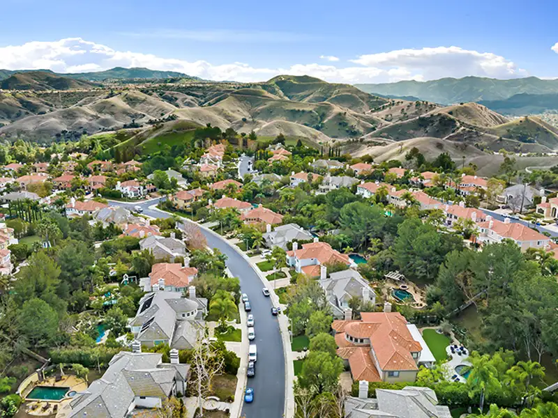 Suburban neighborhood with red-roofed houses and winding road, set against rolling hills under a blue sky.