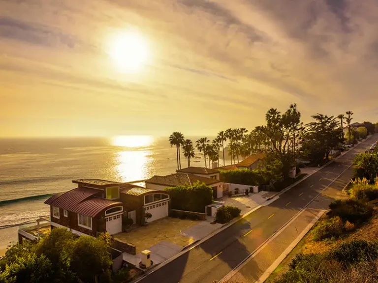 Sunset over oceanfront houses and a palm-lined road.