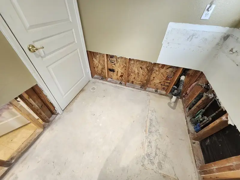 Room with drywall removed from lower walls exposing wooden studs and plumbing pipes.