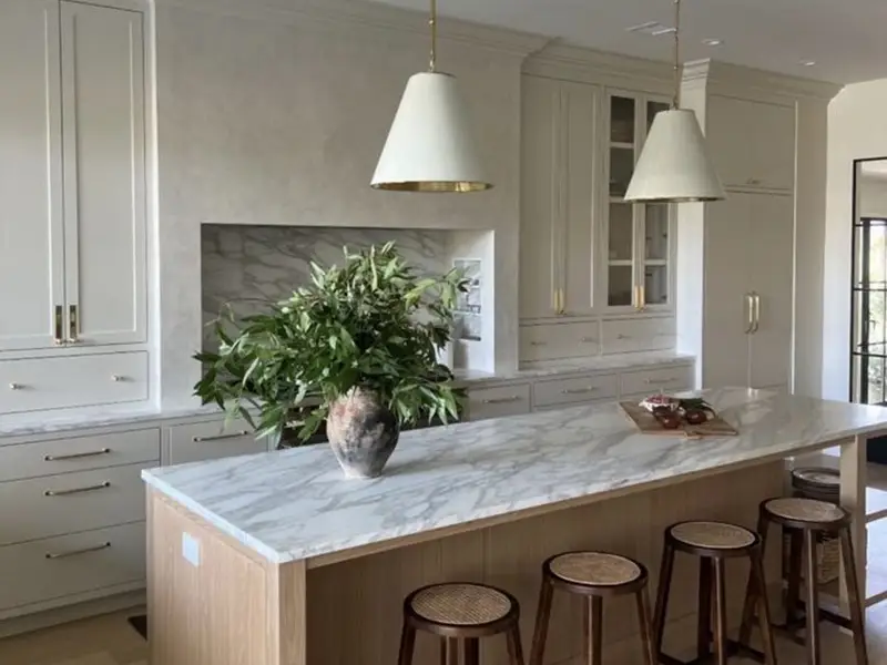 Modern kitchen with marble island, wooden stools, white cabinets, and a vase with green foliage.
