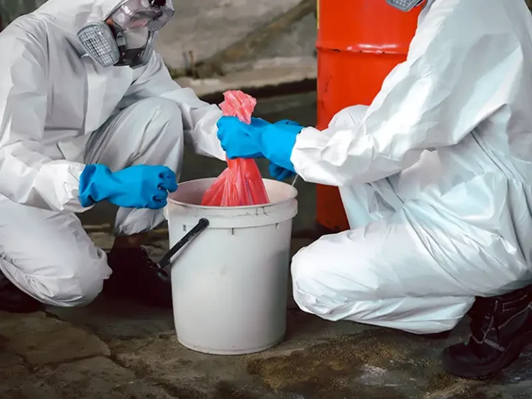 Two people in protective suits and gloves handling a red bag over a white bucket.