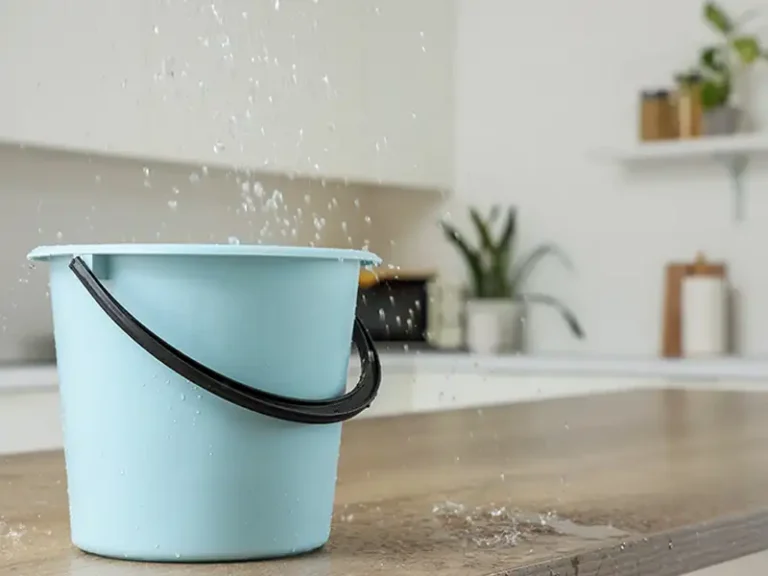 Blue plastic bucket catching water splashes on a wooden countertop in a kitchen.