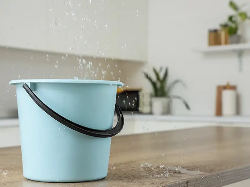 Blue plastic bucket catching water splashes on a wooden countertop in a kitchen.